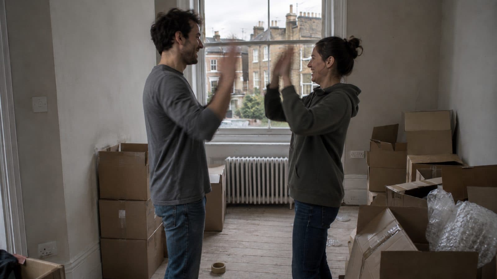 Two people celebrating in a half-unpacked London flat, boxes stacked around them.
