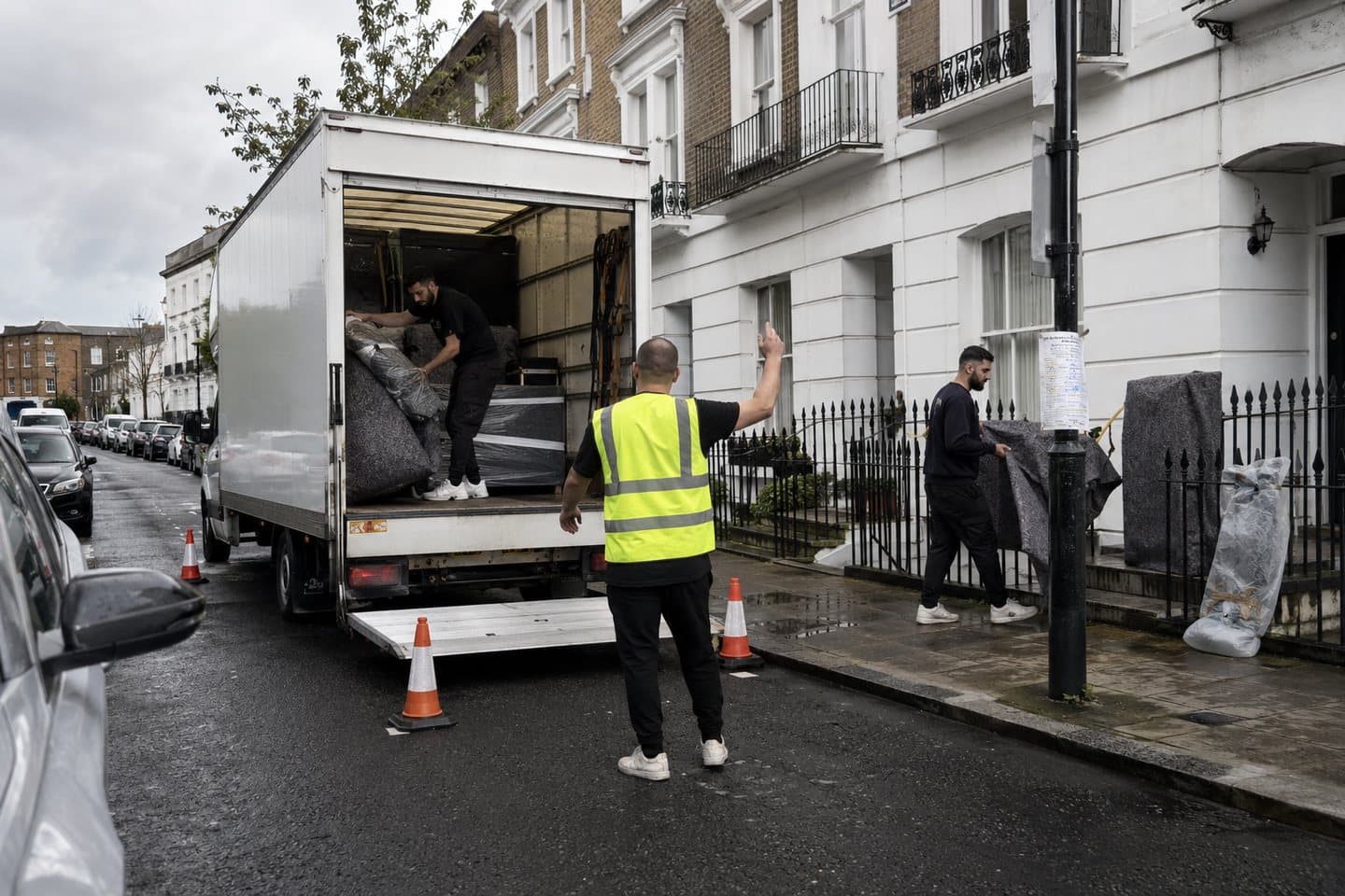 A removals van reversing into a suspended parking bay marked with cones outside a London terraced house, a crew member guiding it back.