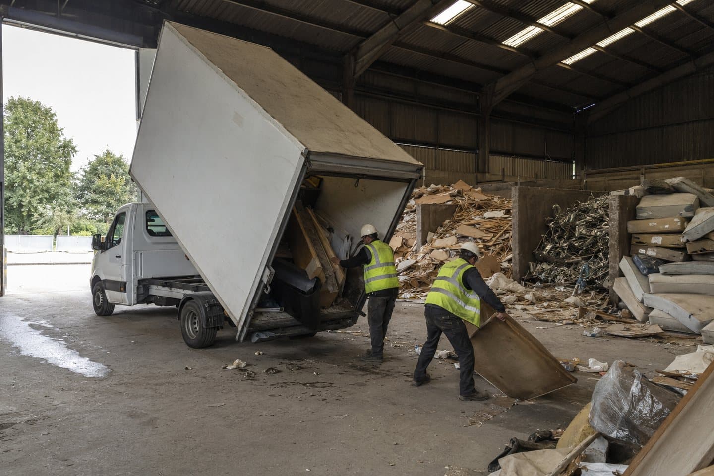A tipper van unloading at a licensed waste transfer station, with separated piles of timber and mattresses and crew in hi-vis and hard hats.