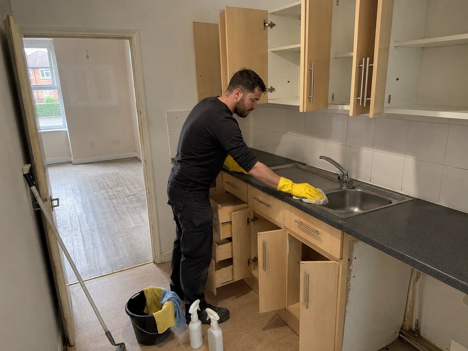 End of tenancy cleaning in London: worker in rubber gloves wiping down the worktop of an emptied rental kitchen, cupboards open and bare