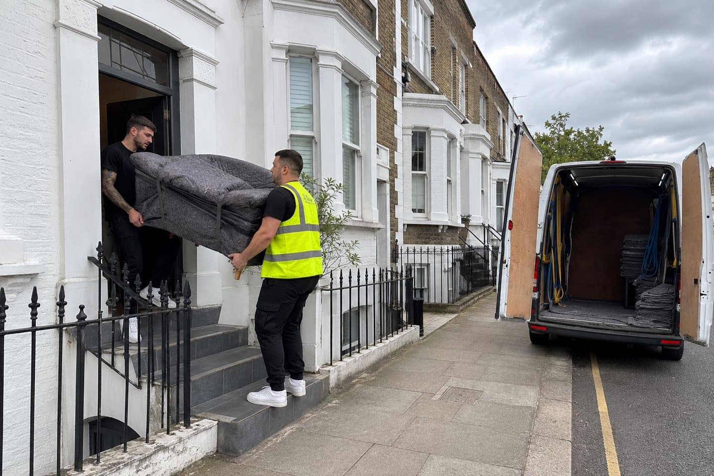 Two removals crew carrying a blanket-wrapped armchair in through the front door of a London terraced house, the van at the kerb behind them.