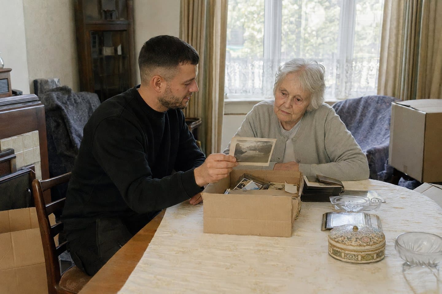A removals crew member and an older woman sitting at a dining table going through a box of photographs together during a house clearance.