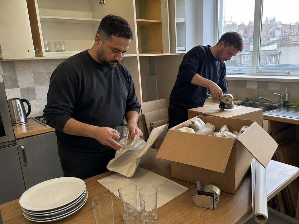Packing services in London: workers wrapping glasses and plates in packing paper at a kitchen table, boxes part filled