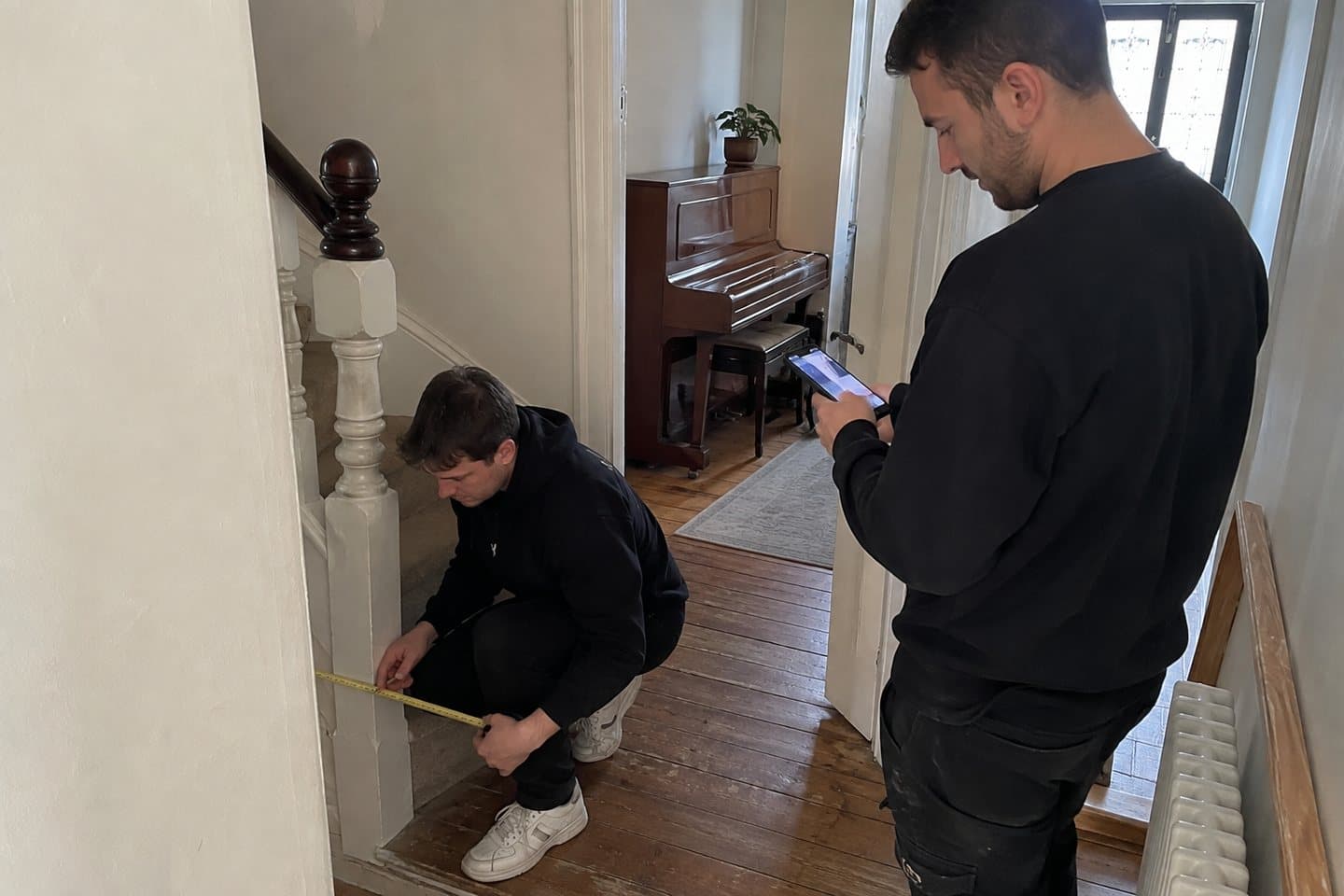A removals crew member measuring the tightest point of a staircase turn with a tape measure, an upright piano visible in the room beyond.
