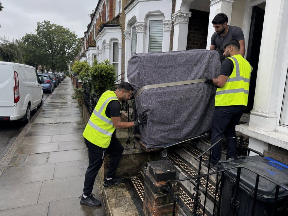 Piano removals in London: Three removal crew easing a blanket-wrapped upright piano down the front steps of a London terraced house on a wheeled piano board